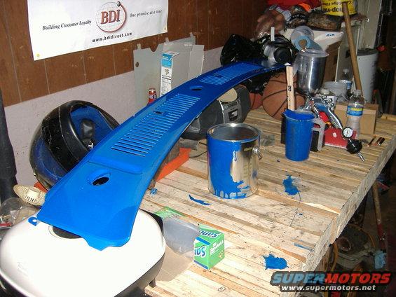 Blue painted car part, possibly a dashboard vent, on a wooden workbench with paint cans and tools
