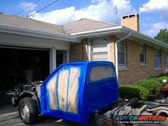 Pickup truck cab with blue paint and masking tape on doors and windows, parked outside a garage