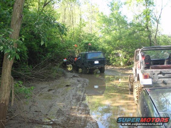 Muddy off-road vehicles navigating a water-filled trail with trees on either side