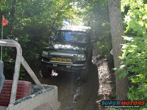 Black 1996 Ford Bronco driving on a muddy trail with trees on either side. Another vehicle is partially visible in the foreground