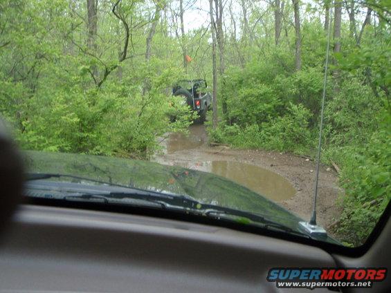 View from inside a vehicle looking at another off-road vehicle navigating a muddy trail in a forest