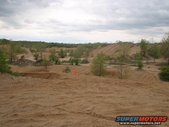 Sandy terrain with tire tracks, trees, and hills under a cloudy sky. An orange cone is visible