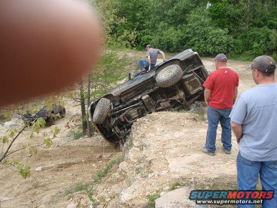 Off-road vehicle overturned on a rocky incline with two people watching. Another vehicle is visible in the background