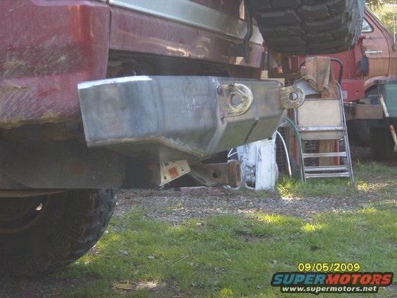 Rear of a red SUV with a custom metal bumper and tow hitch. Spare tire is visible above. Another truck is parked behind it