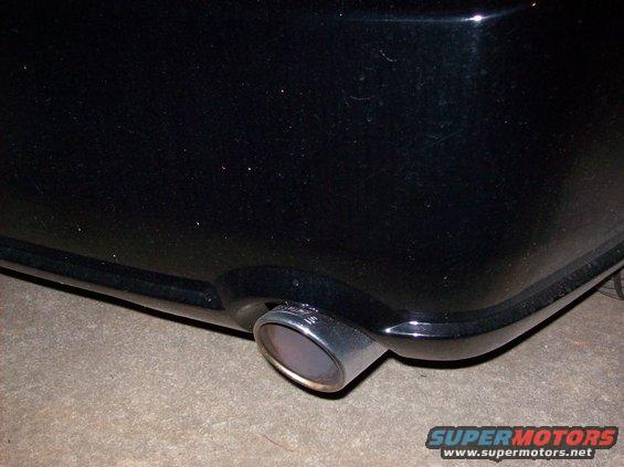 Black car's chrome exhaust pipe visible from the rear quarter panel, with a watermark reading SUPERMOTORS.NET