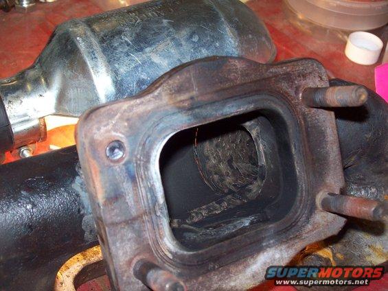 Close-up of a dirty exhaust manifold with a catalytic converter in the background. Text on the converter is partially visible