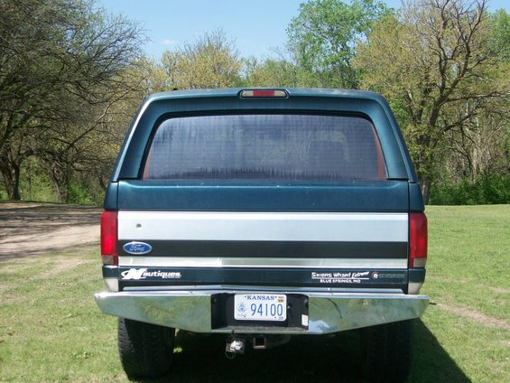 Rear view of a dark green Ford truck with a silver stripe and a camper shell. It has a chrome bumper and a Kansas license plate