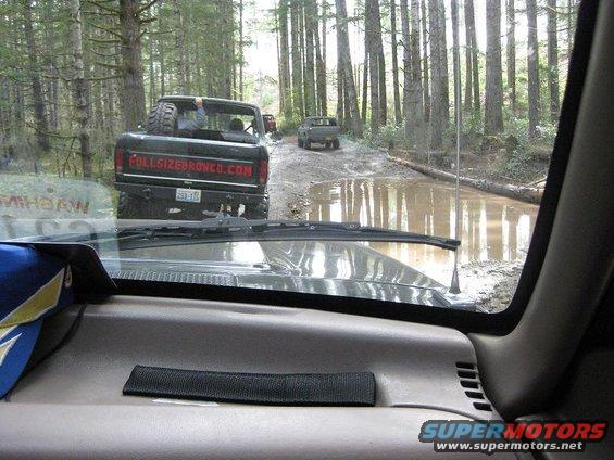 View from inside a vehicle looking at a 1992 Ford Bronco with FULLSIZEBRONCO.COM on the tailgate driving through a muddy forest road