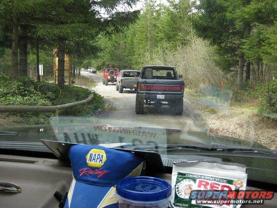 Several trucks driving on a dirt road through a forest. View from inside a vehicle