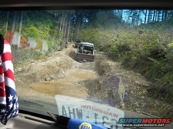 Two off-road vehicles navigate a challenging muddy trail through a forest