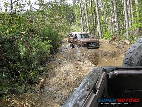 Brown Ford Bronco navigating a muddy trail in a forest, seen from another vehicle