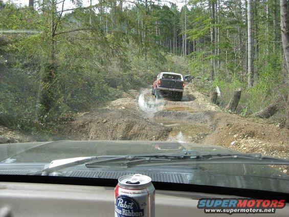 View from inside a vehicle looking at a 1992 Ford Bronco driving on a muddy, wooded trail