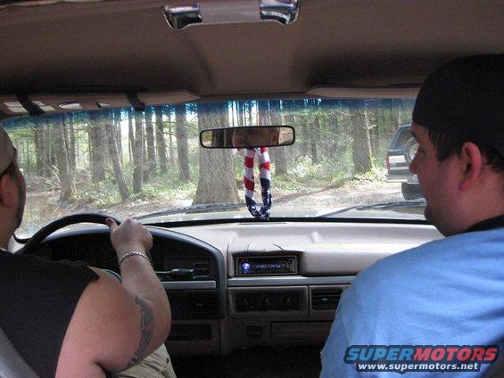 Driving through trees, another SUV ahead. American flag air freshener hanging from mirror