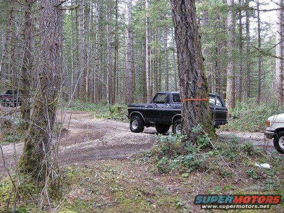 Black Ford Bronco parked on a dirt road in a forest, another vehicle visible to the right