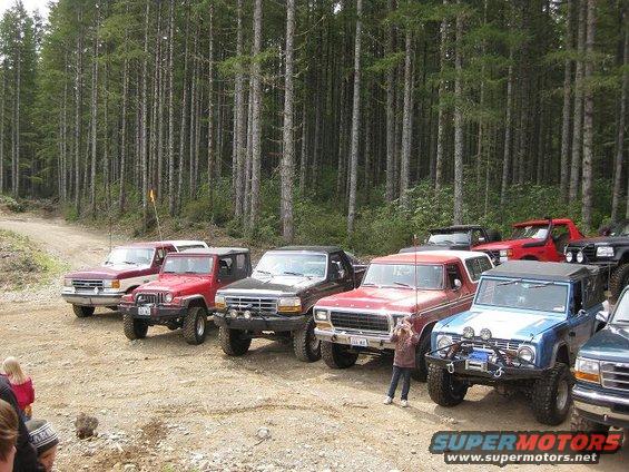 Several off-road trucks and SUVs parked on a dirt road with a forest in the background