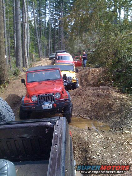 Red Jeep Wrangler driving on a muddy forest trail with other vehicles behind it