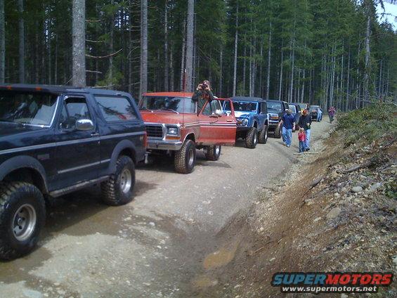 Several off-road vehicles driving down a dirt road in a forest. A red truck has its door open