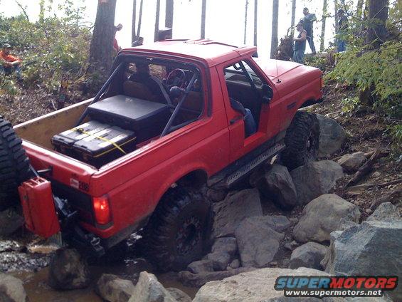 Red 1992 Ford Bronco off-roading over rocks, with a roll cage and extra fuel cans