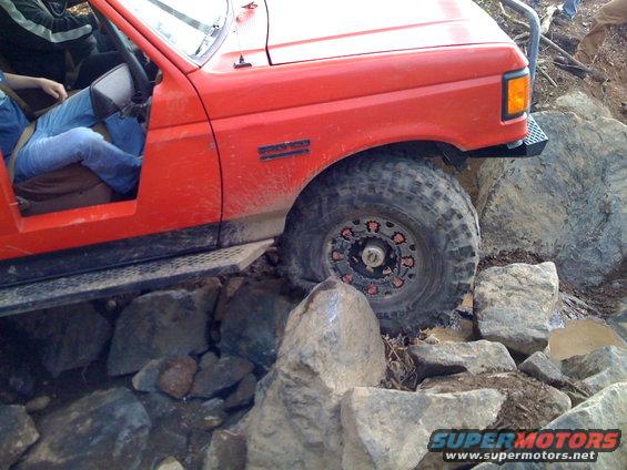 Red SUV with off-road tires navigating large rocks. Driver's door is open, showing a person inside