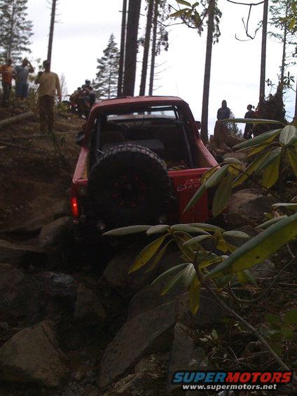 red SUV with spare tire on back driving over rocks on a trail with people watching