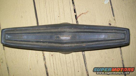 Dark gray car emblem and trim piece with horizontal lines and a central badge, resting on wooden planks