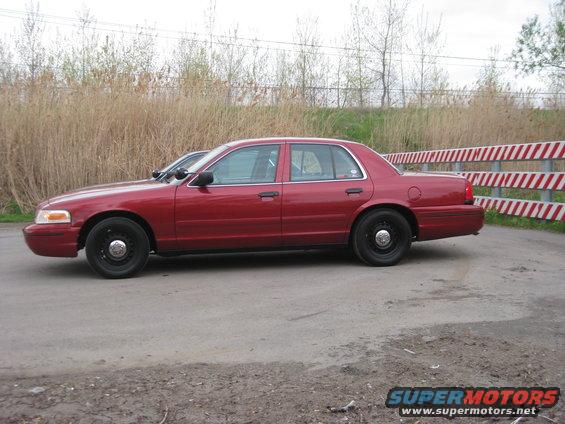 Red sedan parked on a dirt road, another car partially visible behind it. Red and white striped barrier in background