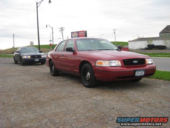 Red Ford Crown Victoria, front 3/4 view, parked on gravel lot with another dark Crown Victoria behind it