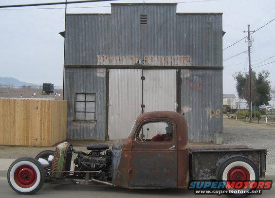 Rat rod pickup truck with exposed engine and red wheels, parked in front of a corrugated metal building