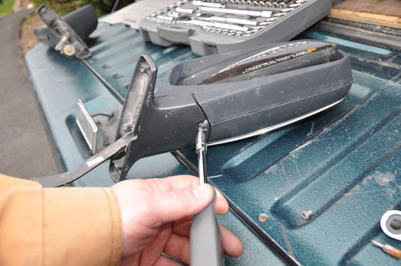 Black car side mirror with chrome trim, being worked on with a screwdriver on a green truck bed