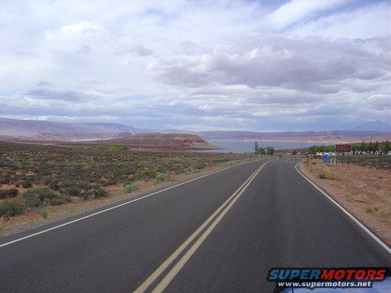 Desert landscape with a two-lane road leading towards a body of water and distant mountains under a cloudy sky