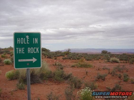 Green sign reading "HOLE IN THE ROCK" with an arrow pointing right, set in a desert landscape under a cloudy sky
