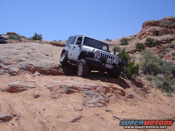 Silver Jeep Wrangler climbing a rocky, sandy trail. Another Jeep is visible in the background