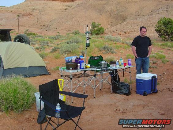 Campsite setup with a tent, folding table with cooking gear, chairs, and a man standing near a cooler