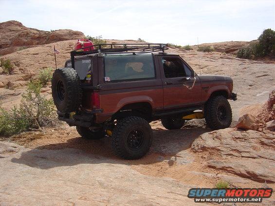 Brown Ford Bronco II with a spare tire on the back, roof rack, and red gas can, driving on a rocky trail