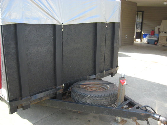 Utility trailer frame with spare tire mounted, black textured walls, and a silver tarp covering the top