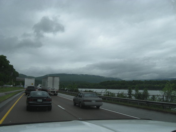Highway traffic scene with trucks and cars under a cloudy sky, with a body of water and hills in the background
