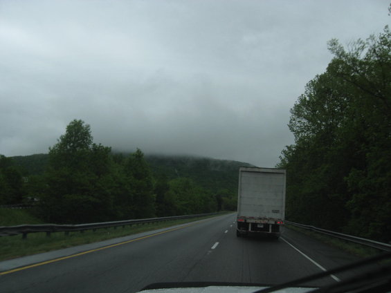 Semi-trailer truck driving on a wet highway with trees and hills in the background under a cloudy sky
