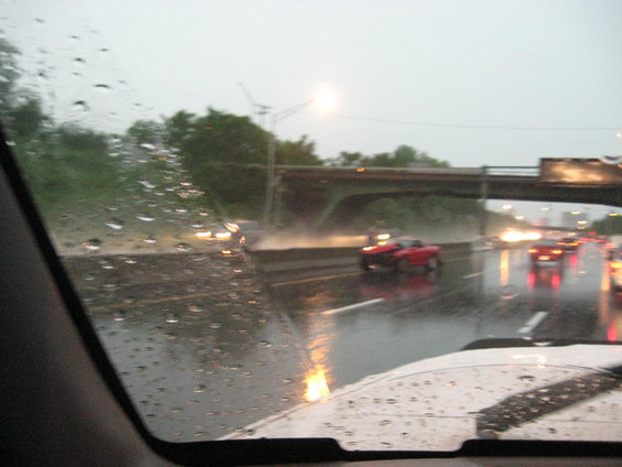 View through a rain-streaked windshield of highway traffic during a downpour, with a red car visible
