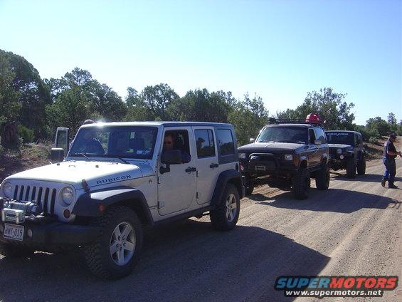 Silver Jeep Wrangler and a dark Ford Bronco II driving on a dirt road with another vehicle behind it