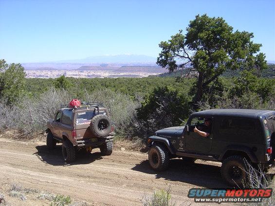 Brown Ford Bronco II and dark green Jeep Wrangler on a dirt road with a scenic overlook