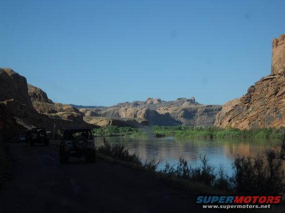 Several off-road vehicles drive on a dirt road next to a wide river with rocky cliffs in the background