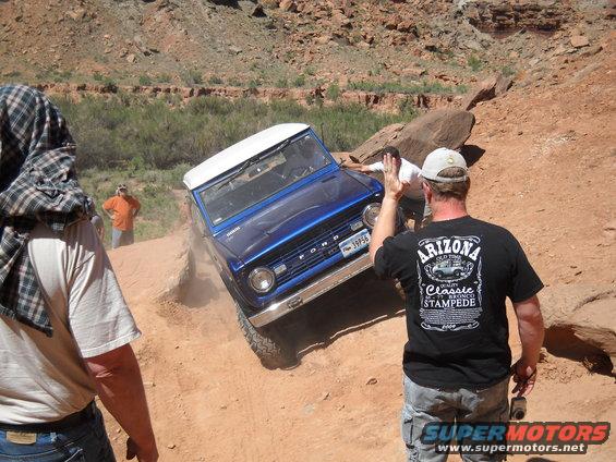 Blue Ford Bronco climbing a dirt hill with people around