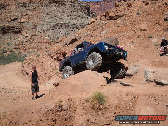 Blue off-road SUV with large tires climbing rocks, front bumper modified, two people visible