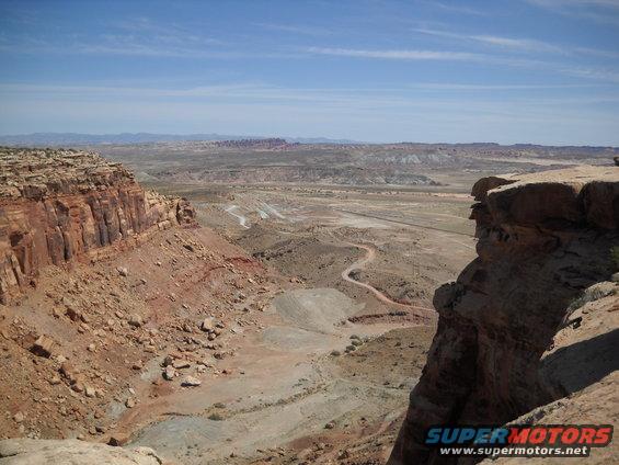Desert landscape with red rock cliffs, a winding dirt road, and distant mountains under a blue sky