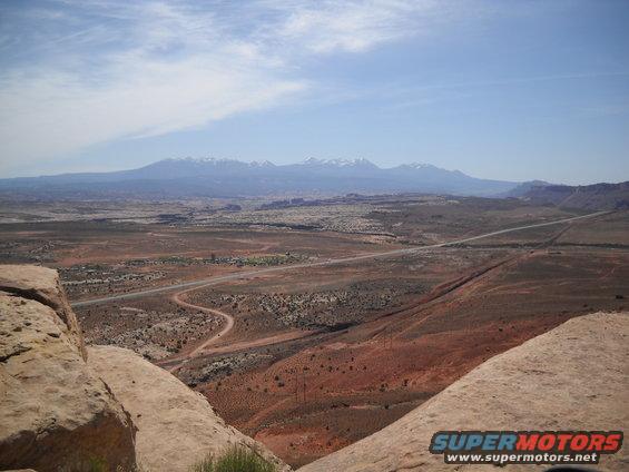 Desert landscape with a winding road and distant mountains under a blue sky. Supermotors logo in the corner