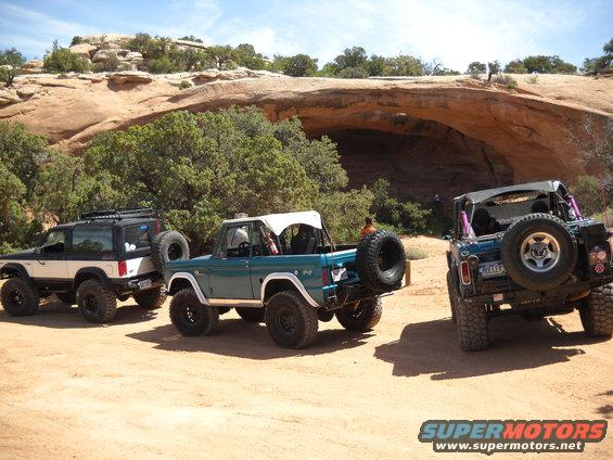 Teal 1978 Ford Bronco, white Ford Bronco II, and blue Ford Bronco parked on dirt with rock formations behind