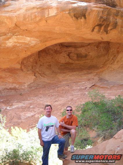 Two men stand in front of a rock overhang in a desert area. A watermark reads SUPERMOTORS