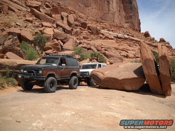 Brown 1986 Ford Bronco II and a white SUV parked on a dirt road with large red rocks and cliffs in the background