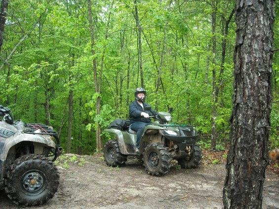 Green ATV with a rider in a helmet, parked on a muddy trail in a forest. Another ATV is partially visible to the left