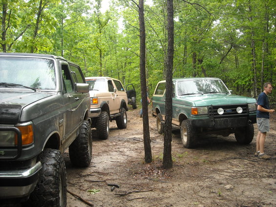 Three lifted Ford Bronco SUVs parked on a dirt path in a forest. One is dark gray, one is tan, and one is green